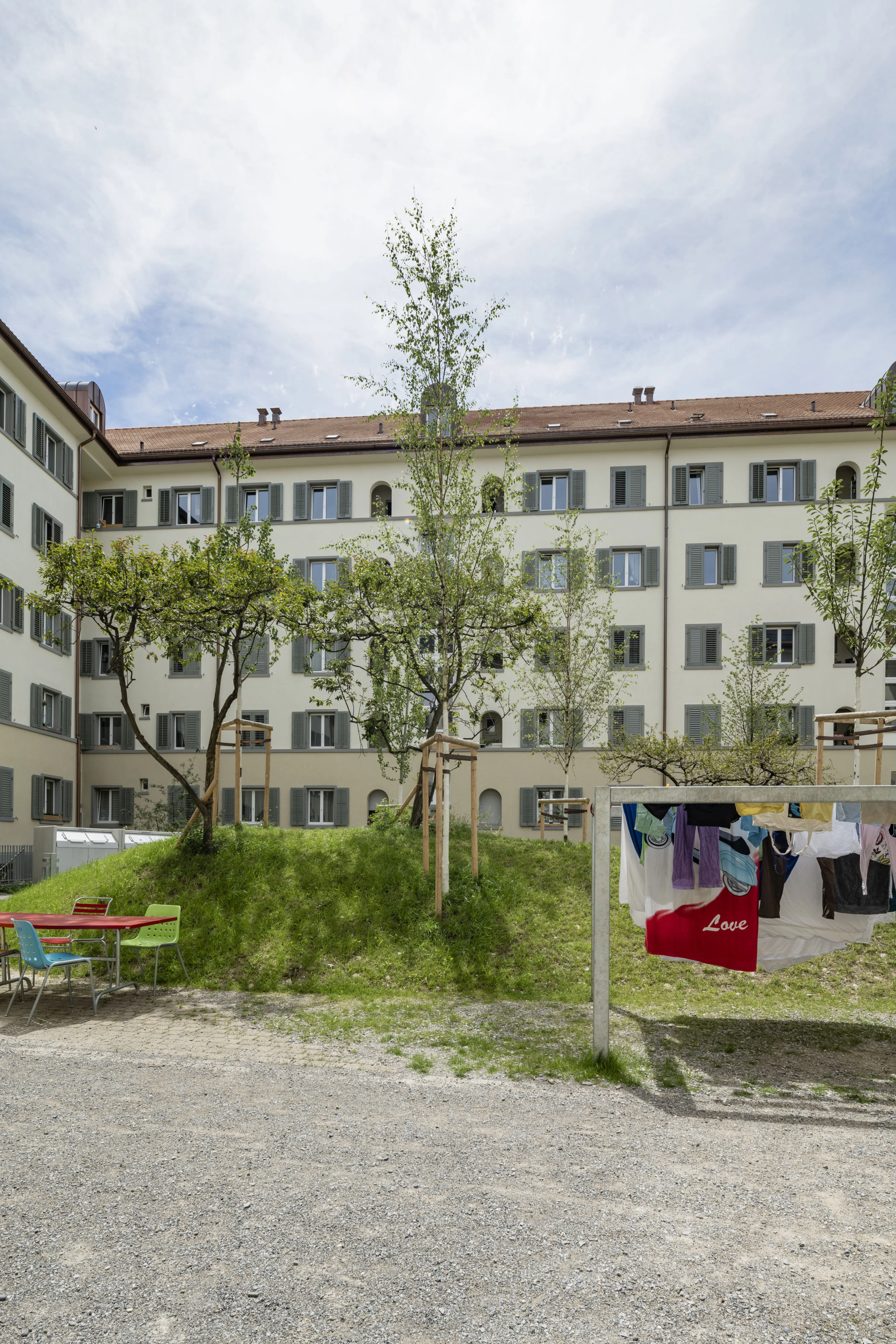 Blick auf die neu modellierte Hügel-Topografie im Innenhof, einen Wäschebereich mit aufgehängter Wäsche und einem roten Tisch mit bunten Stühlen im Vordergrund. Dahinter befinden sich Bäume, Fahrräder und eine helle Fassade eines Wohngebäudes.