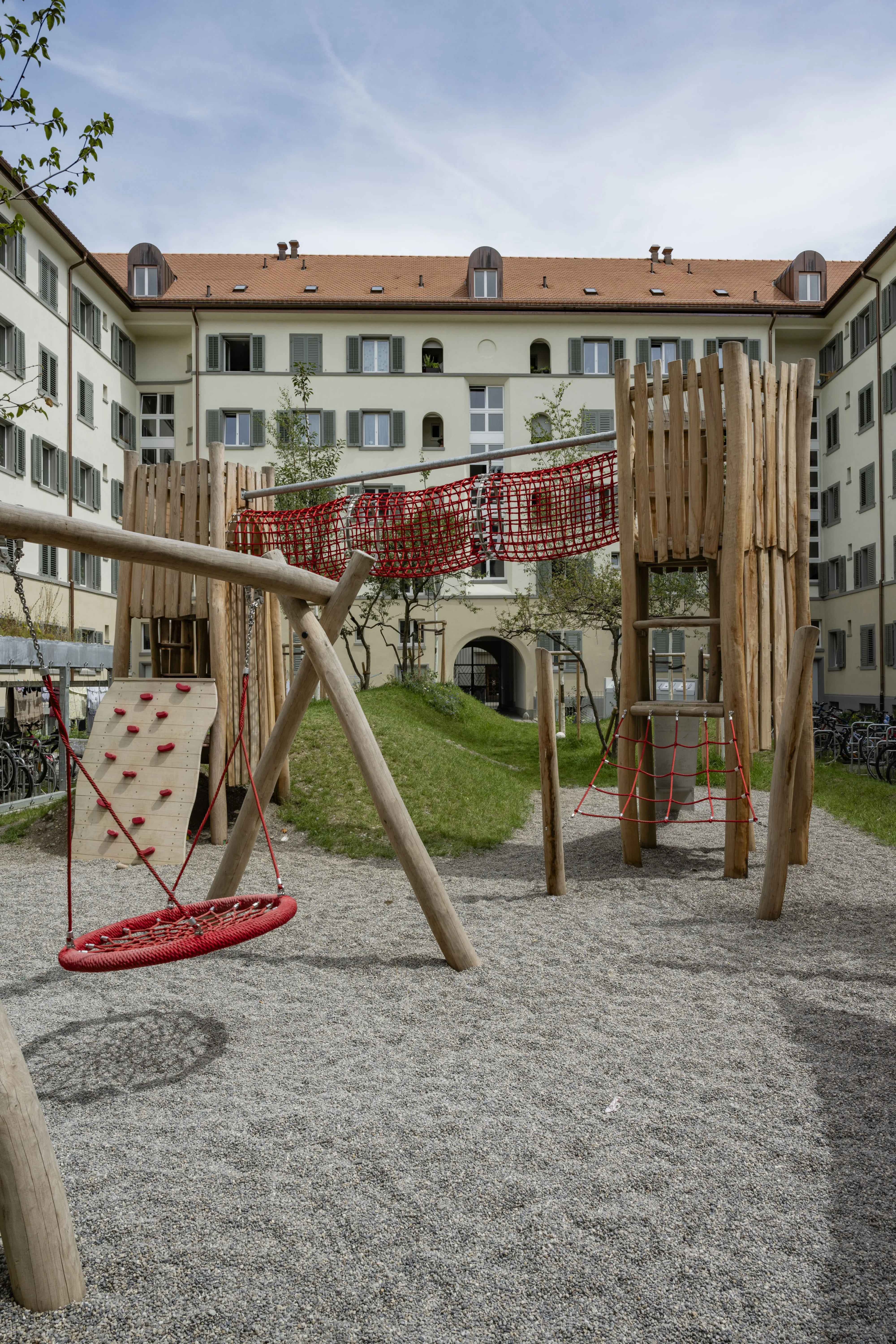 Ein hölzernes Spielgerüst mit roten Netzen und Kletterelementen, aufgestellt im Innenhof einer Wohnsiedlung. Das Wohngebäude im Hintergrund hat hellgelbe Fassaden und mehrere Stockwerke.