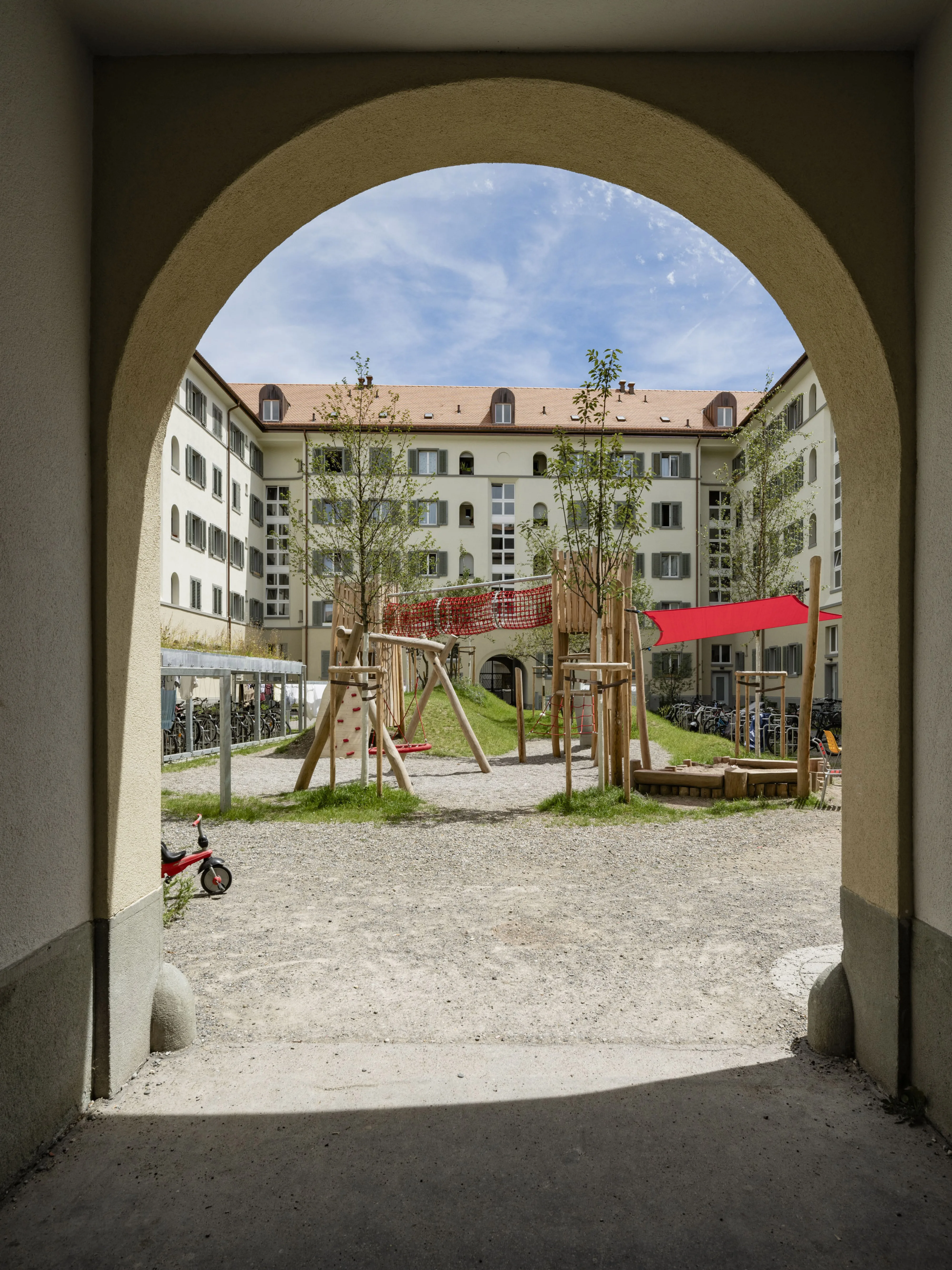 Blick durch den Eingangsbereich in Form eines Torbogen auf den Innenhof mit Spielgeräten aus Holz und roten Kletternetzen. Der Innenhof ist von einem mehrstöckigen Gebäude mit hellen Fassaden und Fensterläden umgeben.