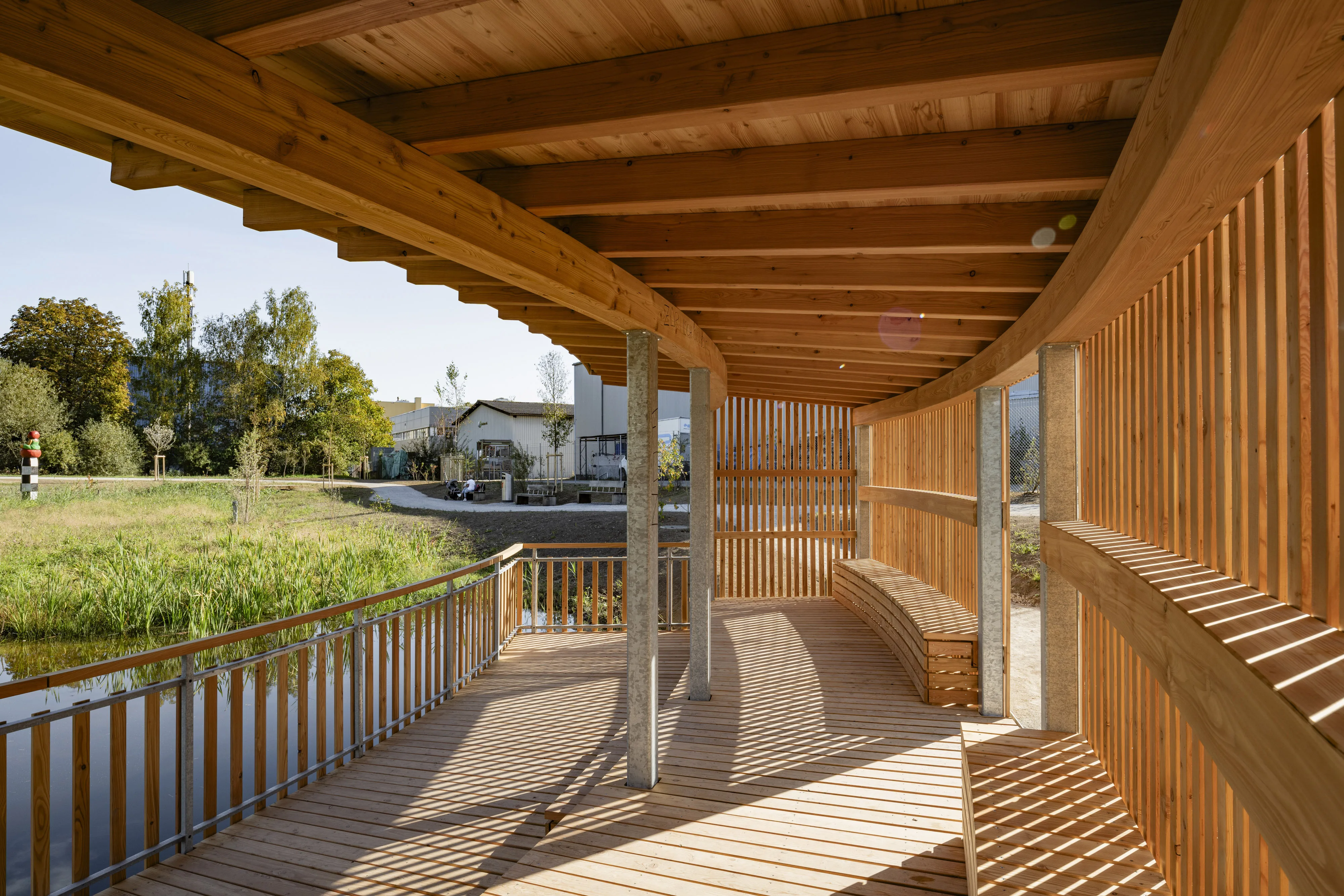 Das Bild zeigt das Innere einer hölzernen Aussichtsplattform, die über einen Teich hinausragt. Die Struktur besteht aus einer offenen, geschwungenen Holzverkleidung und einem überdachten Bereich, der Schatten spendet. Die Licht- und Schattenspiele auf dem Boden und den Wänden betonen das natürliche Design. Im Hintergrund ist eine grüne Wiese und dichte Vegetation zu erkennen, während eine farbenfrohe Skulptur auf einem Pfosten sichtbar ist. Die Plattform bietet eine ruhige Aussicht auf die umliegende Natur und lädt zum Verweilen ein.