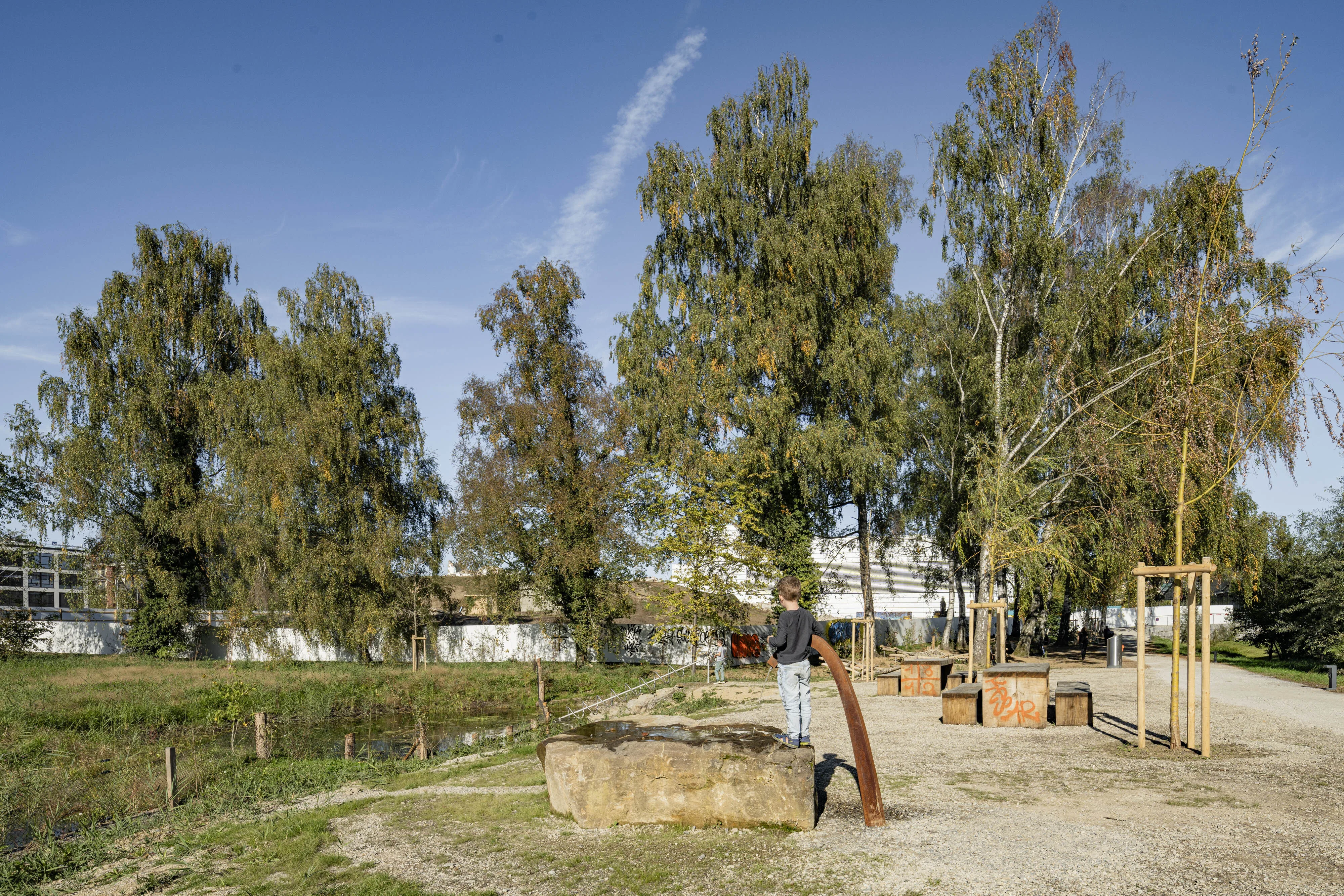 Das Bild zeigt einen jungen Jungen, der auf einem grossen Stein steht und an einem gebogenen Wasserspiel aus Metall spielt, aus dem Wasser fliesst. Der Stein und das Wasserspiel befinden sich auf einem Kiesweg in einer offenen Grünfläche, umgeben von Bäumen und Büschen. Im Hintergrund sind weitere Holzelemente und kleine Teiche zu sehen. Die Bäume im Bild haben ein dichtes, herbstliches Blätterdach, und der Himmel ist klar und blau. Die Szene vermittelt ein naturnahes Spiel- und Erholungsumfeld.