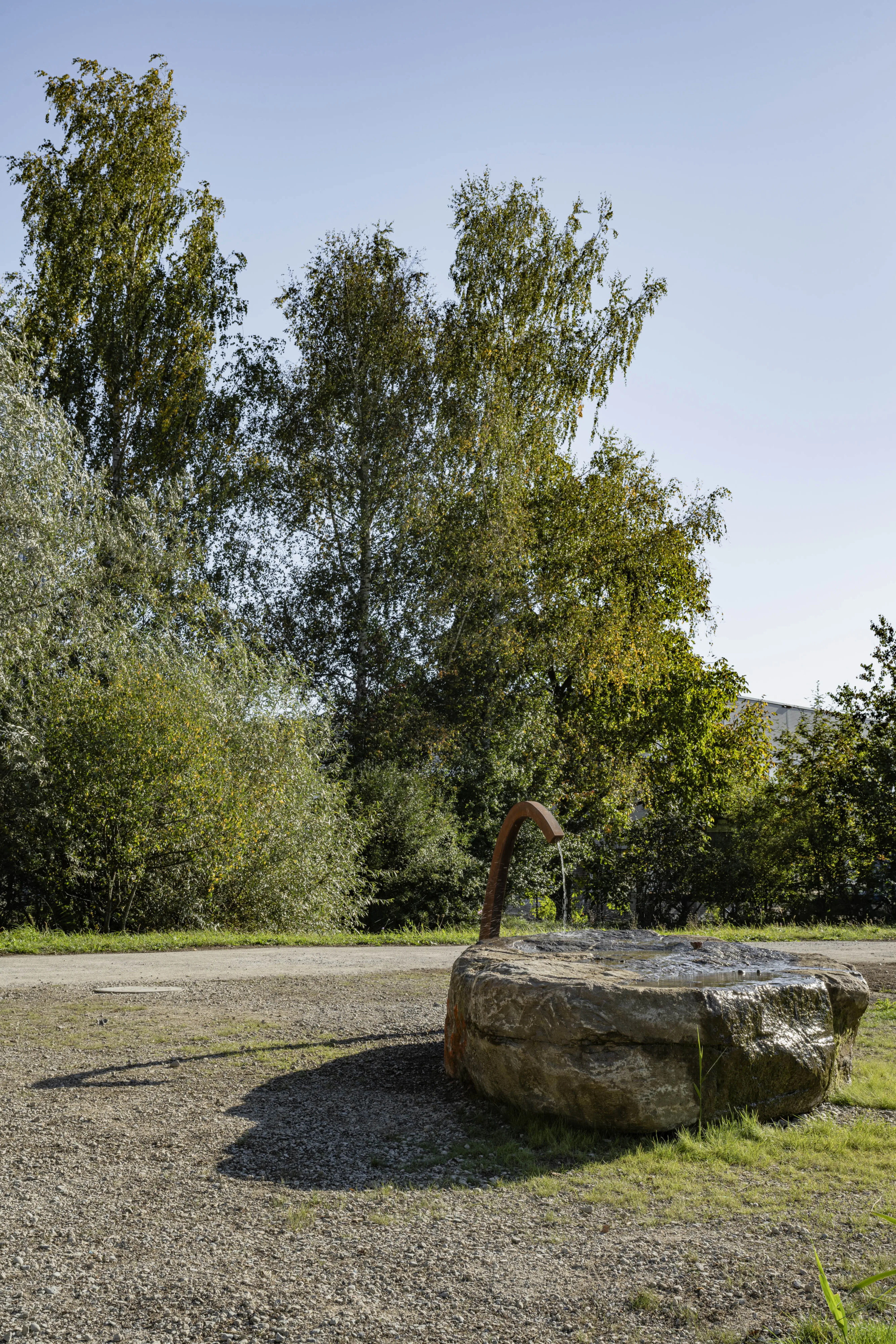 Das Bild zeigt eine rustikale Wasserspielinstallation, bei der ein metallisches Rohr Wasser auf einen grossen, runden Stein fließen lässt. Die Installation befindet sich auf einem Kiesplatz, umgeben von dichtem, grünem Laub und hohen Bäumen im Hintergrund. Die Szenerie wirkt ruhig und naturverbunden, mit einer klaren, blauen Himmel und sanften Schatten, die von den Bäumen und dem Wasserspiel geworfen werden.