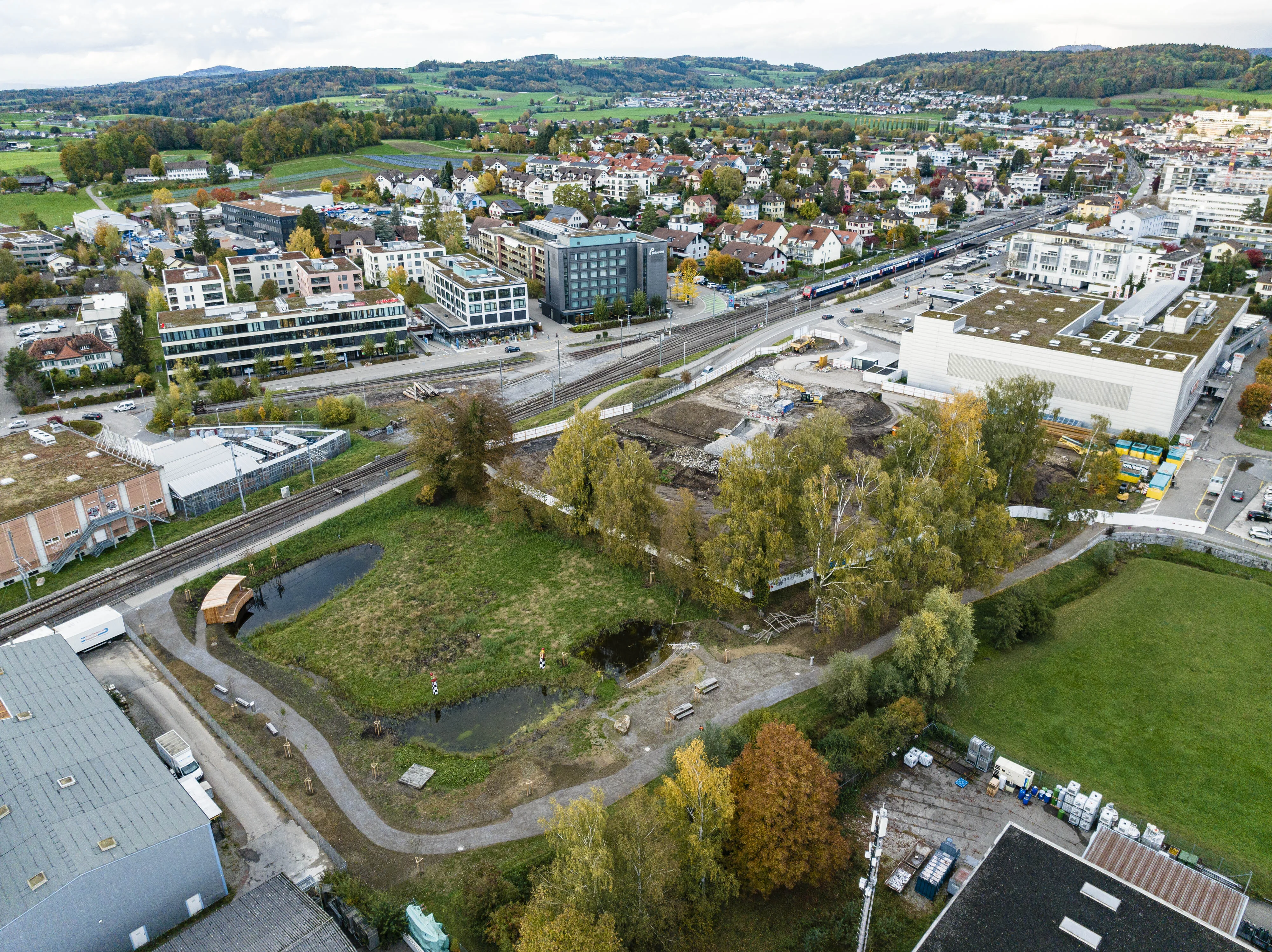 Die Luftaufnahme des Perimeters zeigt eine städtische Landschaft mit einer Mischung aus Gebäuden, Grünflächen und einem kleinen Park. Im Vordergrund befinden sich zwei Teiche, ein geschwungener Weg und eine hölzerne Sitzplattform. Umgeben wird dieser Bereich von Bäumen, darunter auch solche mit herbstlich gefärbtem Laub. Im Hintergrund verlaufen Eisenbahnschienen, an denen moderne Gebäude, Wohnhäuser und Bürokomplexe angrenzen. Weiter hinten erstreckt sich eine hügelige, teils bewaldete Landschaft, die in den Aussenbereich übergeht.